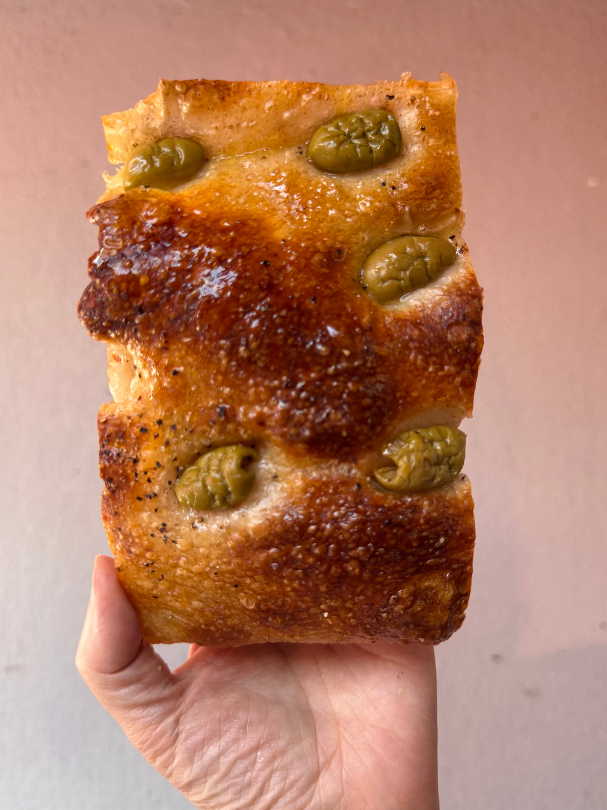 Hand holding a slice of sourdough focaccia with green olives against a neutral background