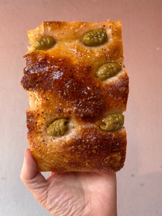Hand holding a slice of sourdough focaccia with green olives against a neutral background